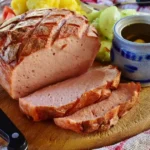 Sliced turkey meatloaf on a wooden cutting board served with mustard, cucumbers, tomatoes, and potatoes.