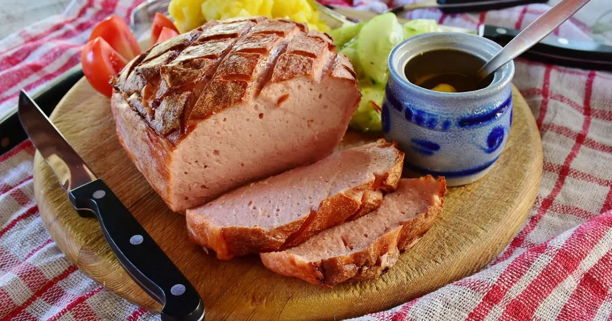 Sliced turkey meatloaf on a wooden cutting board served with mustard, cucumbers, tomatoes, and potatoes.